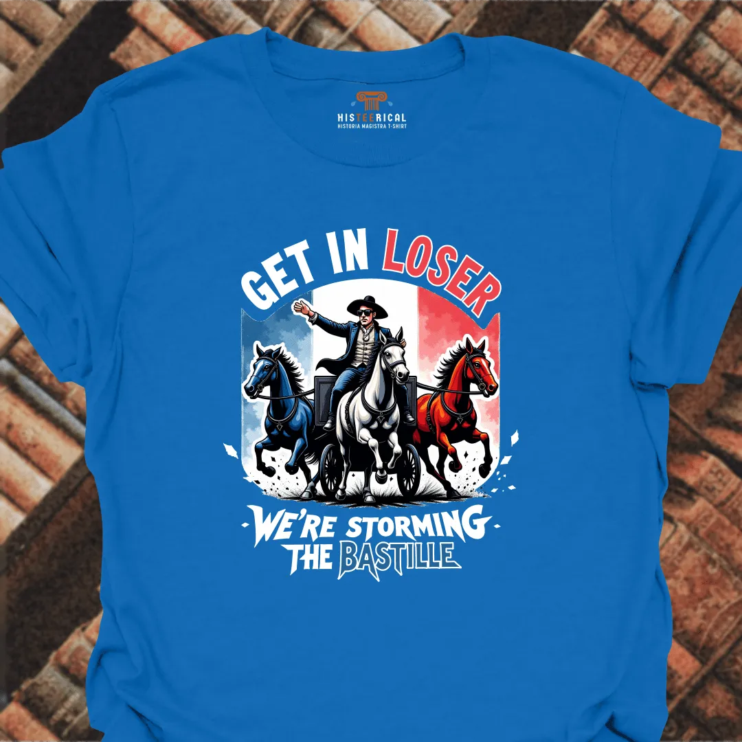 Blue t-shirt with graphic design of a man on horseback and text 'Get in Loser, We're Storming the Bastille' on a brick background.